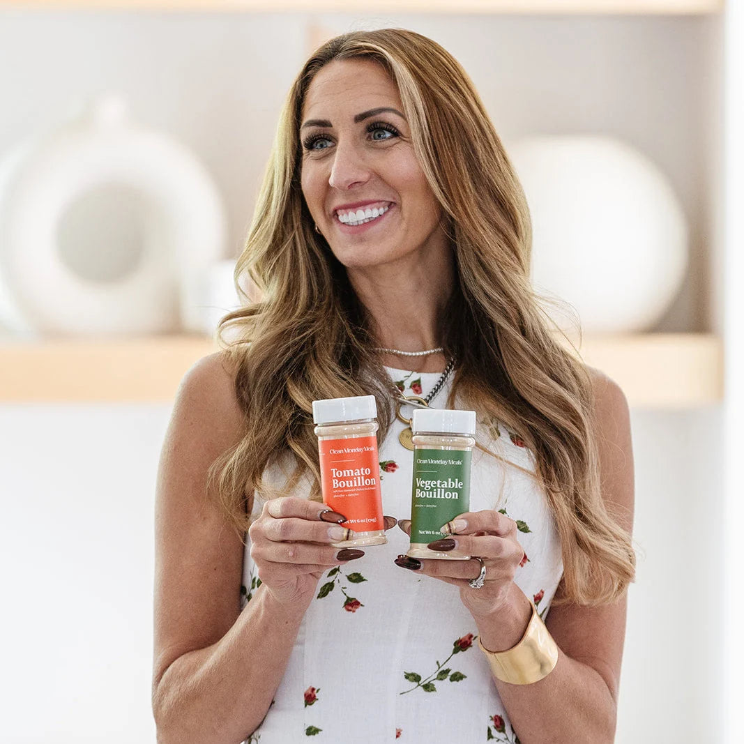 A smiling woman in a floral dress holds two Clean Monday Meals seasoning jars, Tomato Bouillon and Vegetable Bouillon, in a bright kitchen setting.