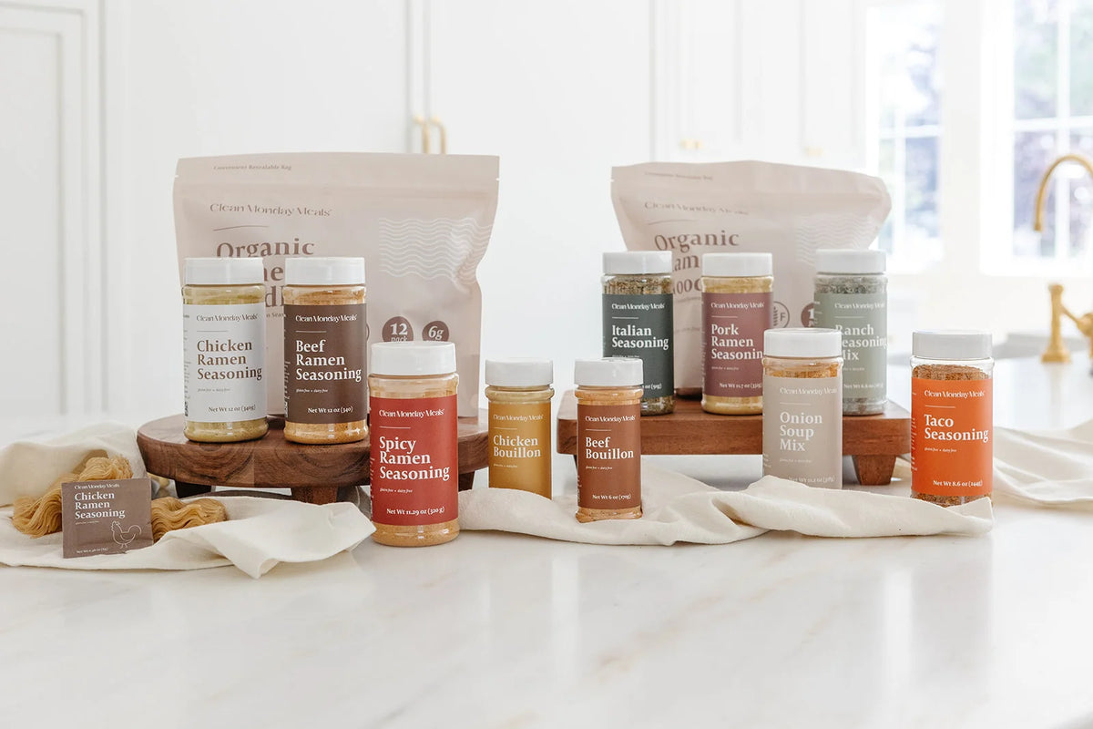 A collection of Clean Monday Meals organic seasoning jars and bags on a white kitchen counter with wooden trays, including Chicken Ramen, Beef Ramen, Spicy Ramen, Italian, Pork Ramen, Ranch, Taco, Onion Soup Mix, Chicken Bouillon, and Beef Bouillon.