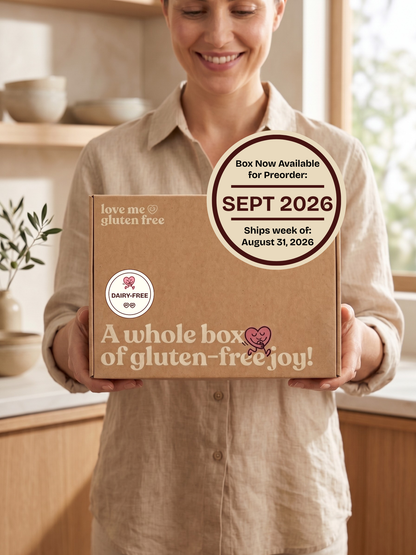 A smiling woman in a beige shirt holds a cardboard box in a bright kitchen setting. The box is brown kraft paper with printed text reading 'love me gluten free' and 'A whole box of gluten-free joy!' plus a circular preorder callout for Sept 2026.