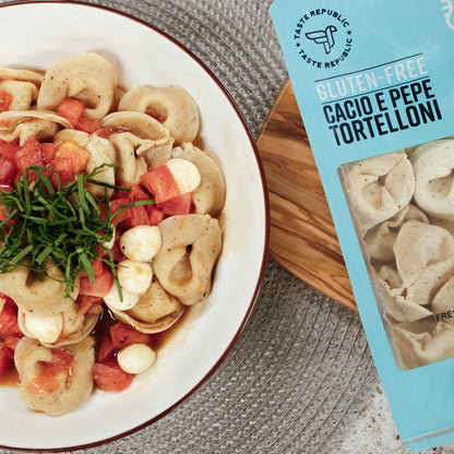 Taste Republic gluten-free Cacio e Pepe tortelloni pasta in a bowl with pesto and caprese ingredients on a wooden table.