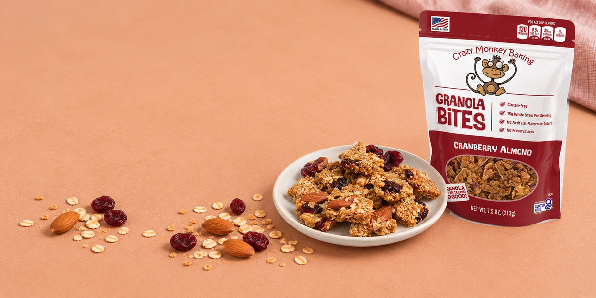 A stand-up pouch of Crazy Monkey Baking Granola Bites Cranberry Almond on a peach background, next to a small white plate of granola bites with scattered oats, almonds, and cranberries.