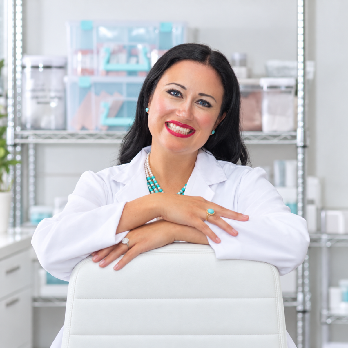 A smiling woman with dark hair, wearing a white lab coat over a turquoise and white patterned top, sitting in a white chair in a bright, modern clinical or laboratory setting with shelving in the background.