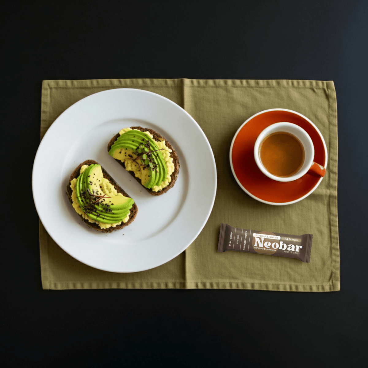 Overhead flat-lay of a white plate with two avocado toast slices, a cup of coffee on an orange saucer, and a Neobar chocolate bar on an olive green placemat against a black background.