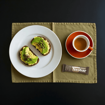 Overhead flat-lay of a white plate with two avocado toast slices, a cup of coffee on an orange saucer, and a Neobar chocolate bar on an olive green placemat against a black background.