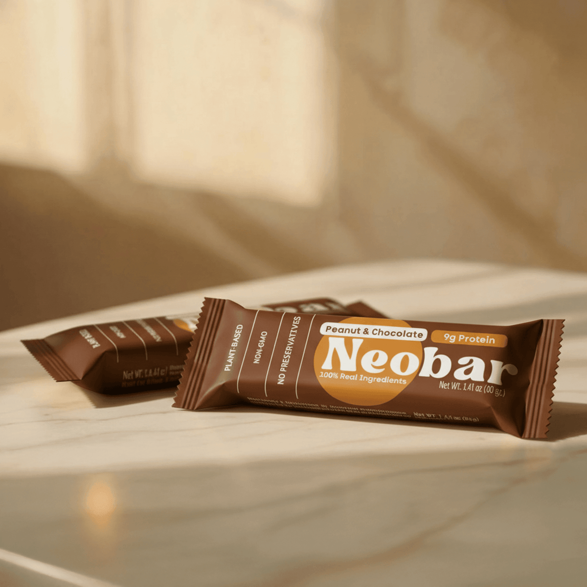 Two Neobar protein bars in brown wrapper, one standing upright and one lying flat, on a light marble surface with warm sunlight and soft shadows.