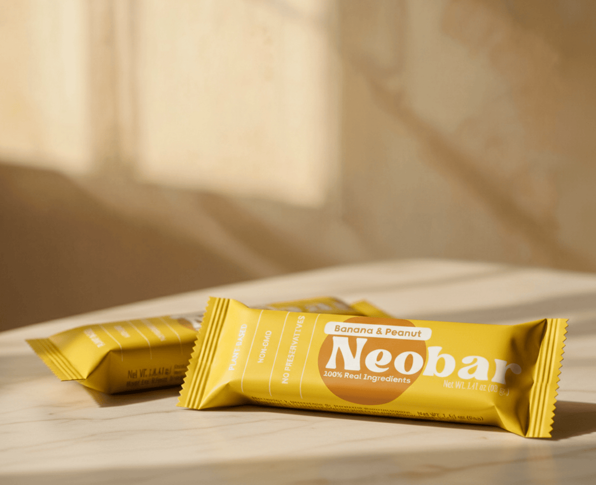 Two yellow Neobar banana and peanut energy bars in foil packaging, arranged on a light marble surface with a warm, sunlit background.