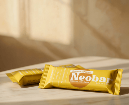 Two yellow Neobar banana and peanut energy bars in foil packaging, arranged on a light marble surface with a warm, sunlit background.
