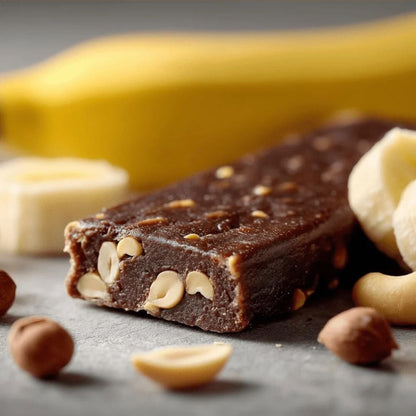 A close-up of a chocolate banana energy bar with visible banana pieces and nuts, photographed on a dark surface with a blurred banana in the background.