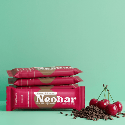 Three Neobar chocolate bars stacked on a mint green background, with red cherries and chocolate chips placed to the right. The packaging is bright pink with white and gold text.